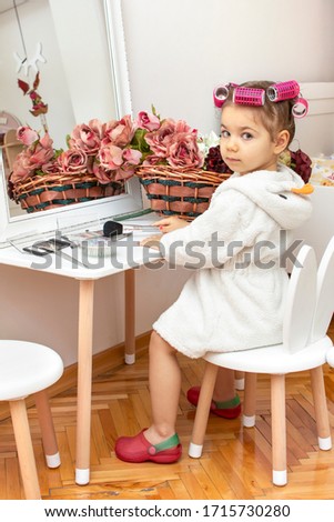 Portrait of happy funny child girl in bathrobe with hair curlers play with mother's cosmetics in front of mirror. Kid's fashion. Mothers day, casual lifestyle photo. Women's day or Birthday background