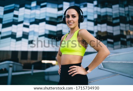 From below of slim athletic female in sports bra and leggings enjoying music in headphones during training while standing on background of glass building and looking at camera