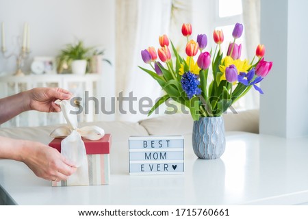 Female hands are opening gift box on marble table with colorful spring flowers bouquet in vase and lightbox with words Best mom ever. Light interior room. Happy Mother's Day background. Copy space