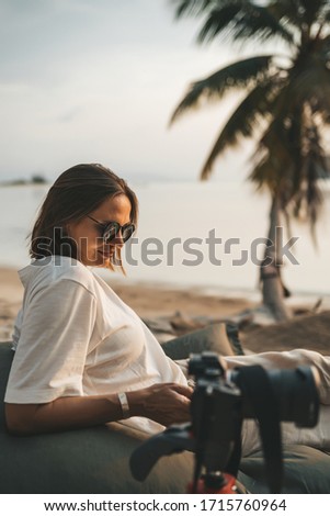 Woman in sunglasses relaxing on beanbag at the beach and making timelapse video with her camera on tripod