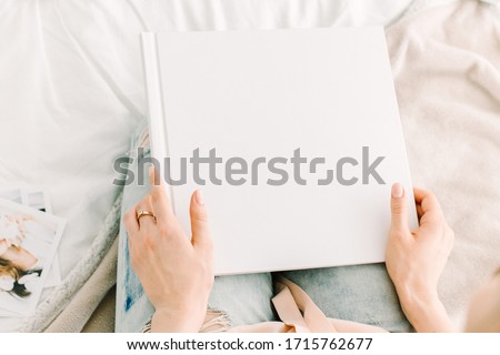 Woman is sitting on the bed with a white cover book mock up in hands. White book template. Square empty book in hands of the woman. Working from home. Office on the bed. Freelancer with a book at home