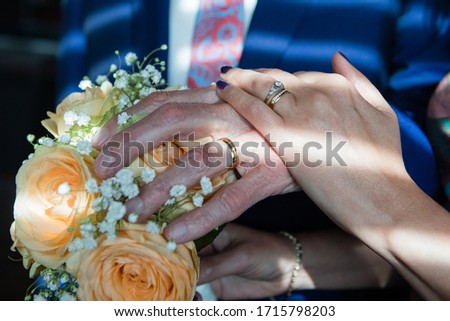 Close up picture of the bride's and elderly groom's hands with yellow gold diamond diamond rings holding together, navy groom's suit, pink tie, wedding bouquet with orange color roses 