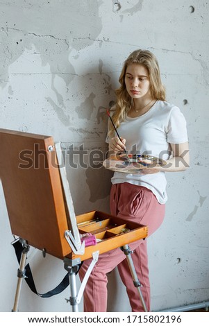 stay home: young sad pretty blonde girl with brush and palette standing near easel drawing picture. Art, creativity, hobby concept, painting process during COVID-19 pandemic self isolation quarantine