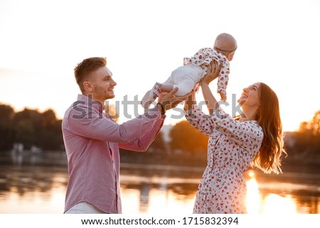 Beautiful woman lifts high her adorable baby girl up mid air and looks at her smiling. Happy parents spending time playing with daughter in park at sunset. Medium shot. selective focus