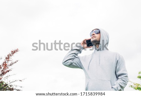 Young man in gray sweatshirt and blue sunglasses holding cell phone in his hands and smiling on a white background