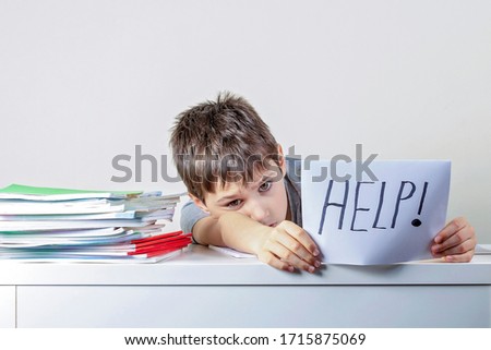 Sad tired frustrated kid sitting at the table with many books and holding paper with word Help. Learning difficulties, education, online learning, school at home