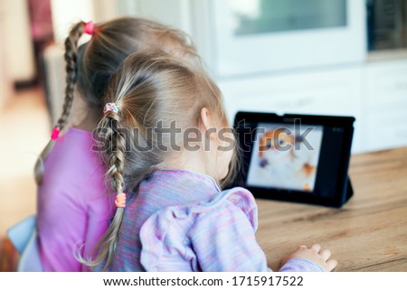  Two girls are sitting at the table and watching videos on the tablet. Learning through the game, learning through the Internet 
