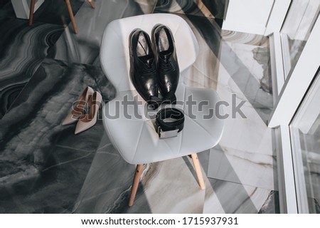 Black shoes, a belt, a groom's bow-tie and cream bride's shoes stand on a chair near the window in a chic interior. Sunlight and shadow. Photography, concept.