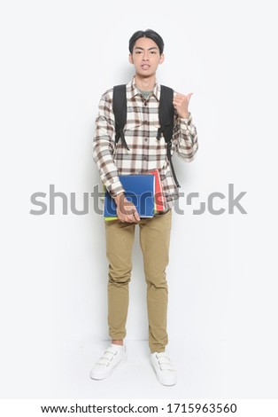 full length cheerful student holding book in hands with standing with backpack making thumbs up sign isolated on white background


