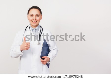Smiling young doctor woman with stethoscope showing thumbs up isolated on white background. Female doctor in medical gown holding health card on notepad folder. Healthcare personnel concept