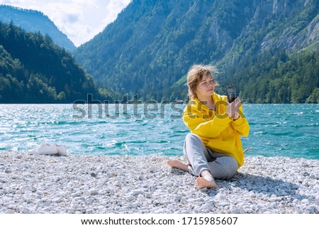 A girl in a bright yellow sweatshirt sits on the shore of a lake in the mountains and takes pictures on a smartphone. Travel concept, people, quarantine, social distance.