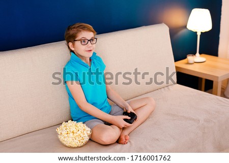 Happy smiling school boy in yellow t-shirt sitting on light couch, playing video games with a gamepad instead of doing his homework at home and eating pop corn. 