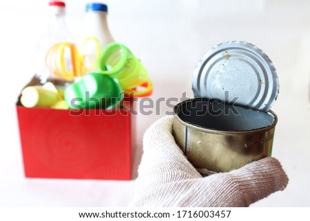 Tin can of canned food in the hand of a man in a glove against the background of a container with plastic waste. The concept of separate waste collection