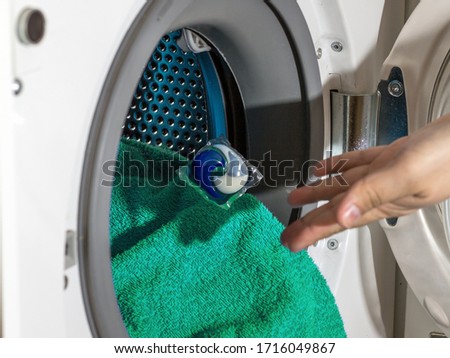 A man throws a washing capsule into a washing machine. The concept of washing clothes and cleanliness. Multi-colored capsule and green towel. Royalty-Free Stock Photo #1716049867
