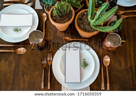 brown wooden table, with white plates, brass cutlery, blank menu sheet and cactus plants