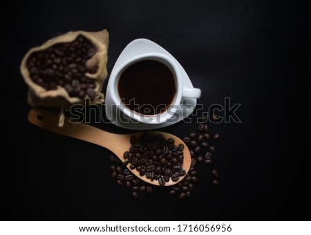 
Roasted coffee beans in small hemp sacks There is a white coffee cup on the side, there is a wooden spoon with coffee beans on top, everything is on a black background.