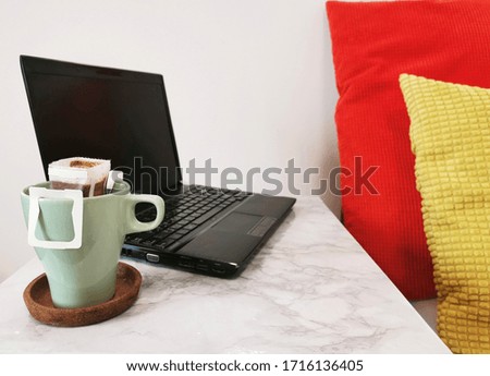 Green drip coffee cup and laptop on marble pattern table with colorful pillow, work from home concept.