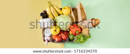 Top view of cardboard box with food products on color green-yellow background. Safe delivery. Contactless home food delivery. Stock for rainy day. Food donation.  Banner with copy space.