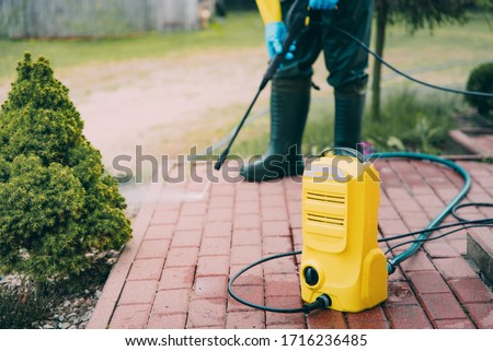 Man cleaning red concrete pavement block using high pressure washer. Paving cleaning concept. Man wearing waders, protective, waterproof trousers doing spring jobs with a jet wash Royalty-Free Stock Photo #1716236485