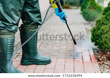Man cleaning red concrete pavement block using high pressure washer. Paving cleaning concept. Man wearing waders, protective, waterproof trousers doing spring jobs with a jet wash Royalty-Free Stock Photo #1716236494