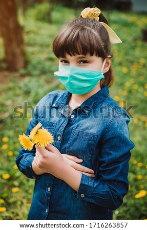 A little girl in denim dress wearing a medical mask, flowers on the background. A child in a protective mask. Warning of the dangers and safety quarantine measures against COVID-19.