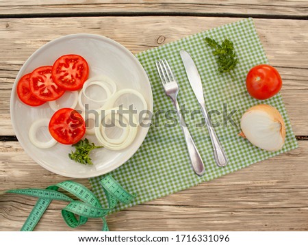 
onion, tomato, parsley, plate with vegetables, knife, fork, measuring tape lie on a cloth napkin on a table in the garden, top view.
sports concept, diet, fitness, plan, healthy eating.