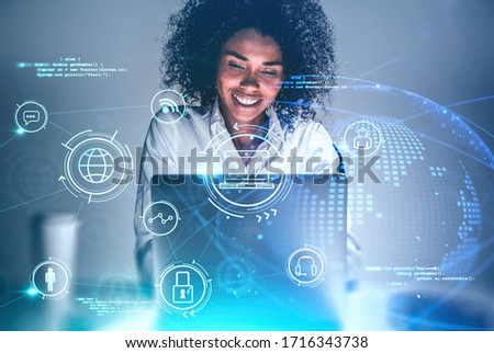 Cheerful young African American woman using laptop at blurry table with double exposure of creative online business interface. Concept of working from home during coronavirus pandemic. Toned image