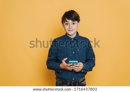 Handsome schoolboy cute smiling, wearing casual shirt and jeans holding his cell phone, looking confident, over yellow backdrop. He got good news.