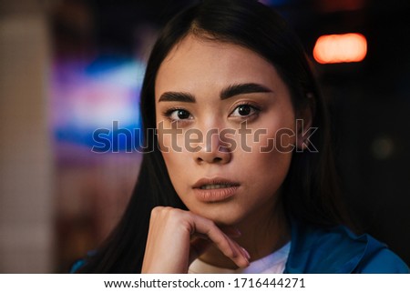 Photo of serious young asian woman with brunette hair looking at camera while posing in cafe