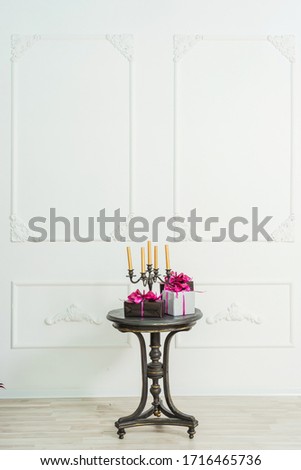 Golden candles in a candelabrum on a black table with gifts boxes on a background of a white wall with stucco molding. Gift boxes in black and silver wrapping paper with pink bows with ribbons