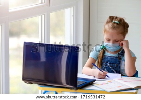 Girl schoolgirl does online homework lessons. Internet school, distance learning, quarantine. Little girl in a medical mask writes in a notebook

