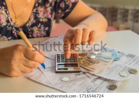 an elderly woman considers payments for utility bills; on a calculator, she considers payment for an apartment with money in Russian rubles; banknotes of 2000,100 rubles and Russian coins Royalty-Free Stock Photo #1716546724