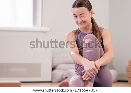 Positive young caucasian athlete who loves pilates and yoga resting after home exercises and sitting on floor looking at camera smiling and winking with one eye. Concept of quarantine home workouts