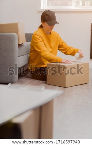 young courier girl seals a cardboard box with duct tape on a light background, vertical photo