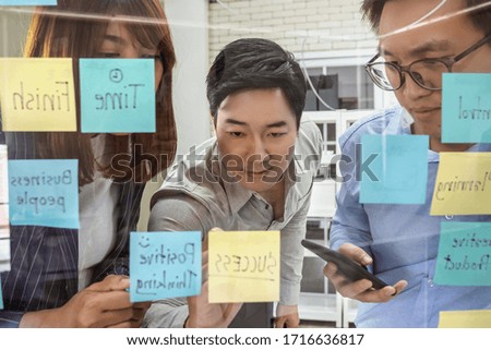 Group of Asian business people working and brainstorming with colleagues together, writing solution with paper notes over the glass wall in office, startup team work and small business concept