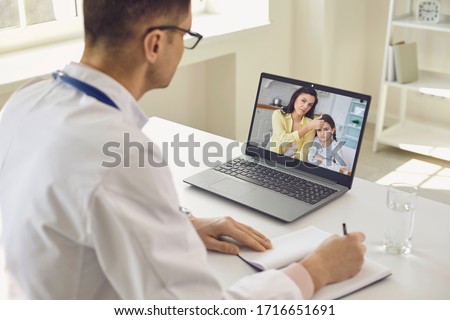Family doctor online. Male doctor speaks with patients by family using laptop sitting behind straw in clinic office. Royalty-Free Stock Photo #1716651691
