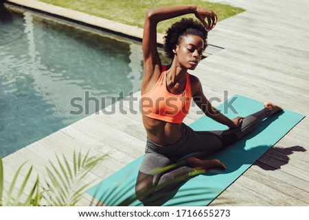 Healthy woman doing yoga stretches by the poolside. African female in sportswear exercising yoga in morning by the swimming pool Royalty-Free Stock Photo #1716655273
