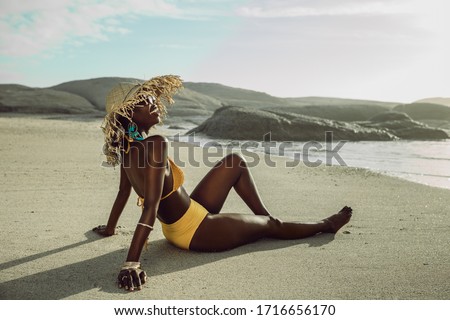 Smiling woman in bikini with sun hat sitting at the beach. African female relaxing on the beach on a summer day.