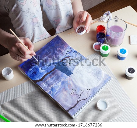 Female hands hold a brush and a cover of a watercolor jar. using it as a palette against the background of a cat and night sky on a white sheet of an album, working tools. Hobbies on Quarantine.