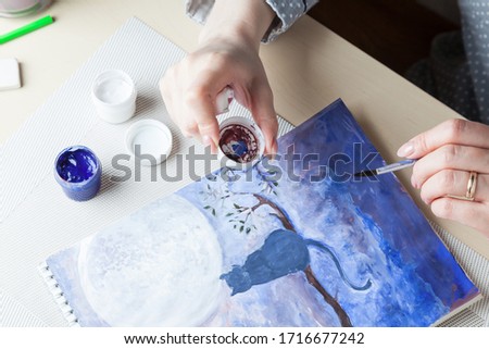 Girl holds in her hands a brush and a cover from a watercolor jar the background of a drawing of a cat, the night sky and the moon made in blue tones, nearby tools. Occupation in quarantine mode.