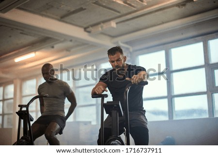 Two fit young men riding stationary bikes together during a cardio workout session at the gym