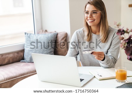 Photo of smiling woman in eyeglasses using laptop while holding credit card at living room