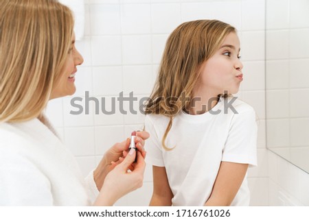 Photo of joyful caucasian mother and daughter smiling while doing makeup in white bathroom