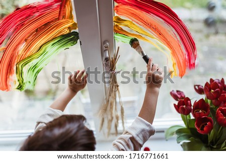 Little boy painting rainbow on a window and looking through the window while sitting home during quarantine. Dirty hands. Let's all be well.
