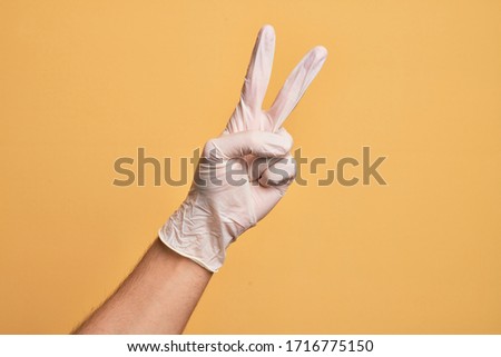 Hand of caucasian young man with medical glove over isolated yellow background counting number 2 showing two fingers, gesturing victory and winner symbol