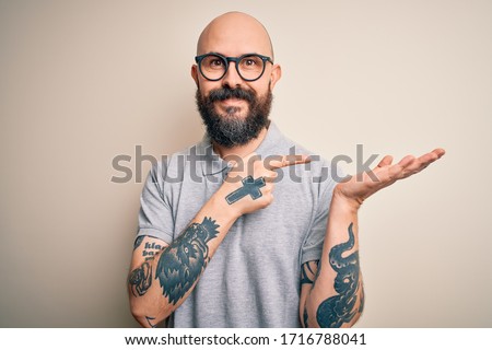 Handsome bald man with beard and tattoo wearing casual polo and glasses amazed and smiling to the camera while presenting with hand and pointing with finger.
