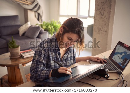 Young woman working from home due to the global pandemic. Illustrator working on the drawing tablet in her living room. Over-Shoulder Shot, smile, medium shop, concept work and artist Royalty-Free Stock Photo #1716803278