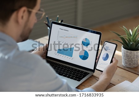 Close up businessman working with statistics, looking at laptop and phone screens with graphs and diagrams, synchronizing electronic devices, checking financial report presentation, sitting at desk Royalty-Free Stock Photo #1716833935