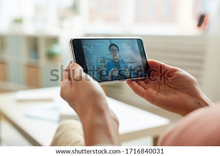 Unrecognizable person watching Asian man speaking while having online meeting on smartphone, horizontal close-up shot Royalty-Free Stock Photo #1716840031