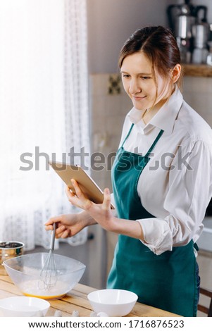 Woman looking to laptop and reading recipe for cooking dishes. Cooking at home concept, lifestyle. Preparing the dough,  baking in the kitchen. Vertical picture